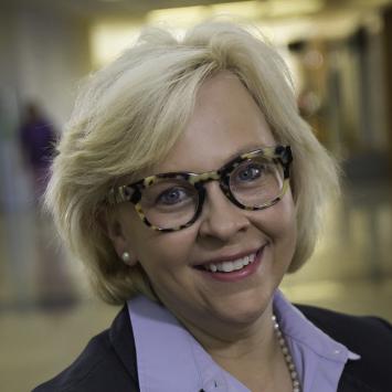 A professional headshot of Julie Balog. She is smiling and wearing tortoiseshell glasses with a purple collared shirt and a suit jacket.