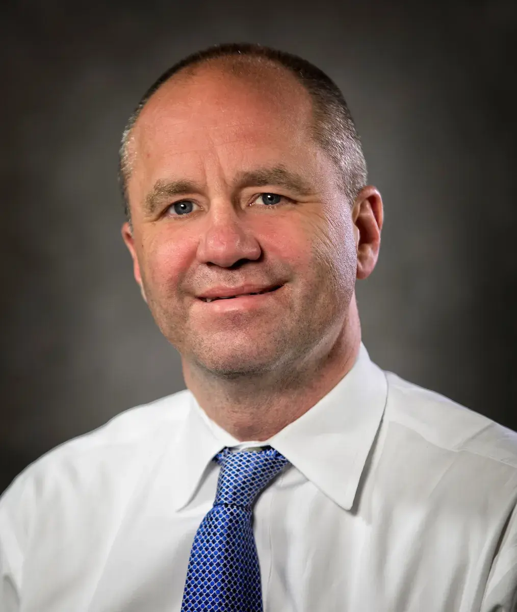 a headshot of Jay Blanton smiling wearing a white collared shirt and a blue tie. The background is a gradient of grey.