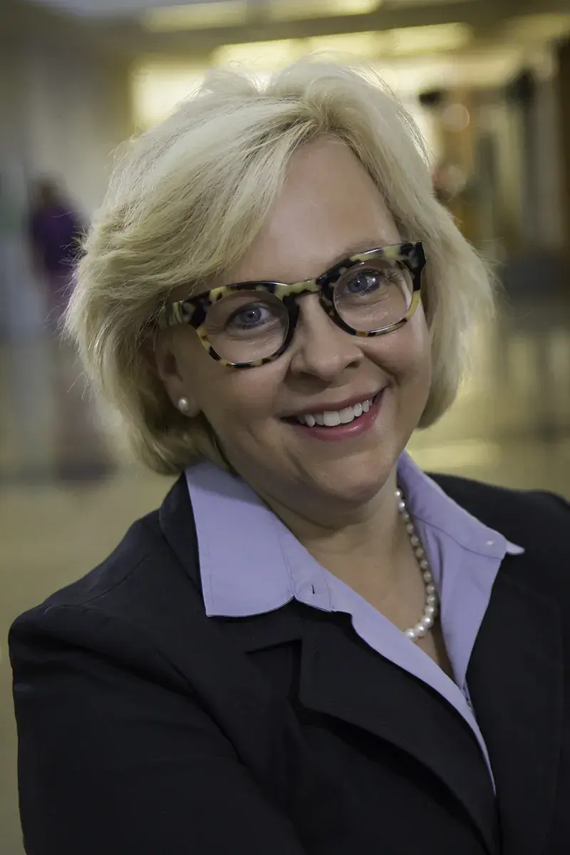 A professional headshot of Julie Balog. She is smiling and wearing tortoiseshell glasses with a purple collared shirt and a suit jacket.